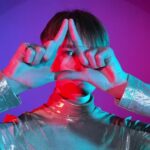 Portrait young adult woman showing masonic symbol, Illuminati triangle symbol hand gesture in neon illumination indoors. Studio shot in art colorful lighting. Female short hair wearing sparkling dress